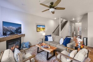 A living room with a fireplace, a television, a sofa, a coffee table, and a ceiling fan. - Big Sky Townhome | Fireplace + Hot Tub (Big Sky)