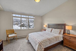 A bedroom with a large window, a bed, a nightstand, a chair, and a lamp. - Big Sky Townhome | Fireplace + Hot Tub (Big Sky)
