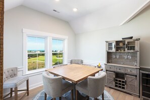 A dining area with a wooden table and four chairs, a kitchen with a built-in wine rack, and a view of a golf course through large windows. - Ocean-view 4-bedroom, 3.5 bath Corrolla home w/pool+hot tub - sleeps 11 (Corolla)