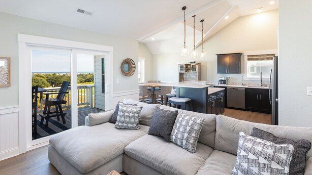 A modern living room with a large sectional sofa, a dining area, and a kitchen with dark cabinets. - Ocean-view 4-bedroom, 3.5 bath Corrolla home w/pool+hot tub - sleeps 11 (Corolla)