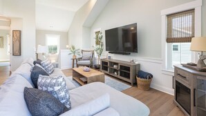 A spacious living room with a high ceiling, a large flat-screen TV mounted on the wall, a long sofa with patterned cushions, a wooden coffee table, and a fireplace with a mantel. - Ocean-view 4-bedroom, 3.5 bath Corrolla home w/pool+hot tub - sleeps 11 (Corolla)