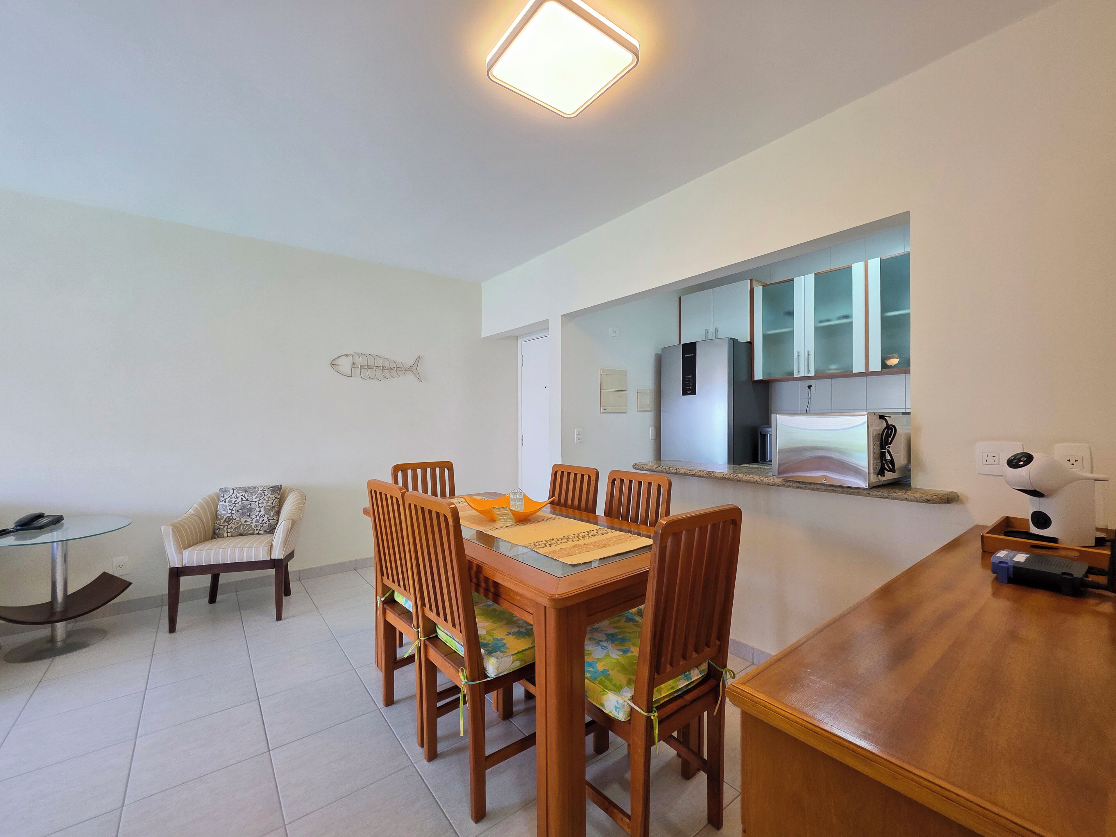 A dining area with a wooden table and chairs, a fish-shaped wall decoration, and a kitchen area with cabinets and a refrigerator.