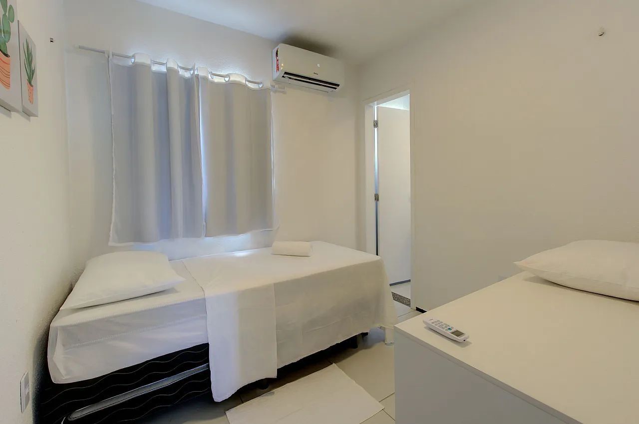 A small, clean room with two beds, a window with curtains, an air conditioning unit, and a remote control on the nightstand.