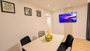 Un coin repas comprenant une table, des chaises, un panier de fruits, des œuvres d’art encadrées et un téléviseur fixé au mur.