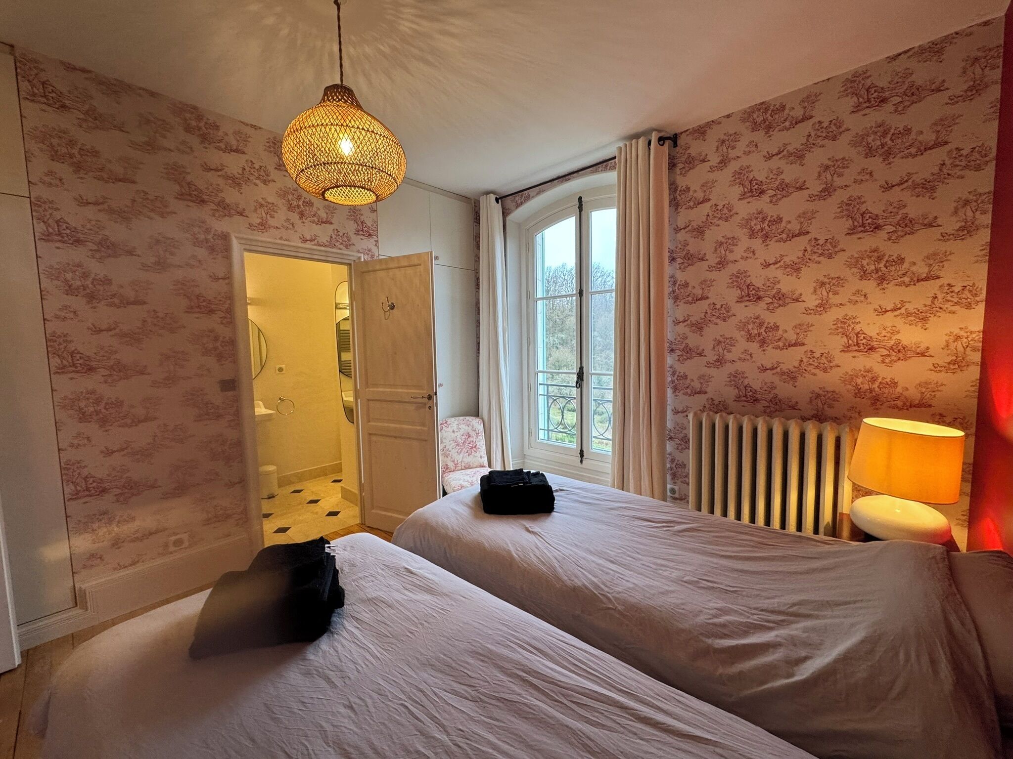 A bedroom with two beds, a hanging light fixture, a window with curtains, and a radiator.