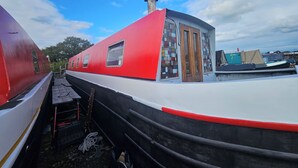 Een rode en witte boot met een klein huisje en schoorsteen, afgemeerd in een jachthaven met andere boten.