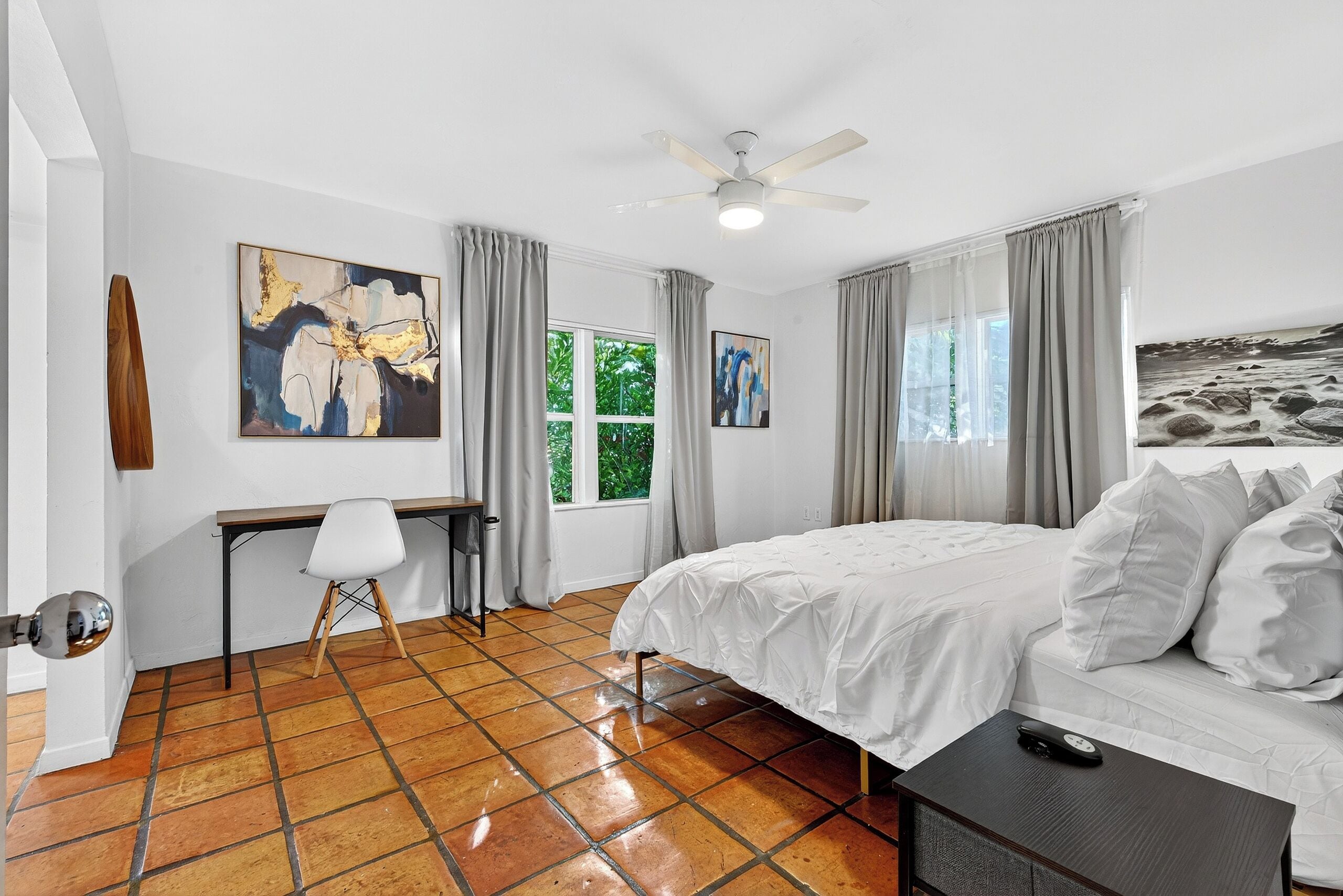 A bedroom with a bed, a desk, and a chair. There are curtains on the windows, and artwork on the walls.