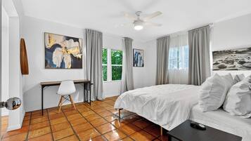 A bedroom with a bed, a desk, and a chair. There are curtains on the windows, and artwork on the walls.