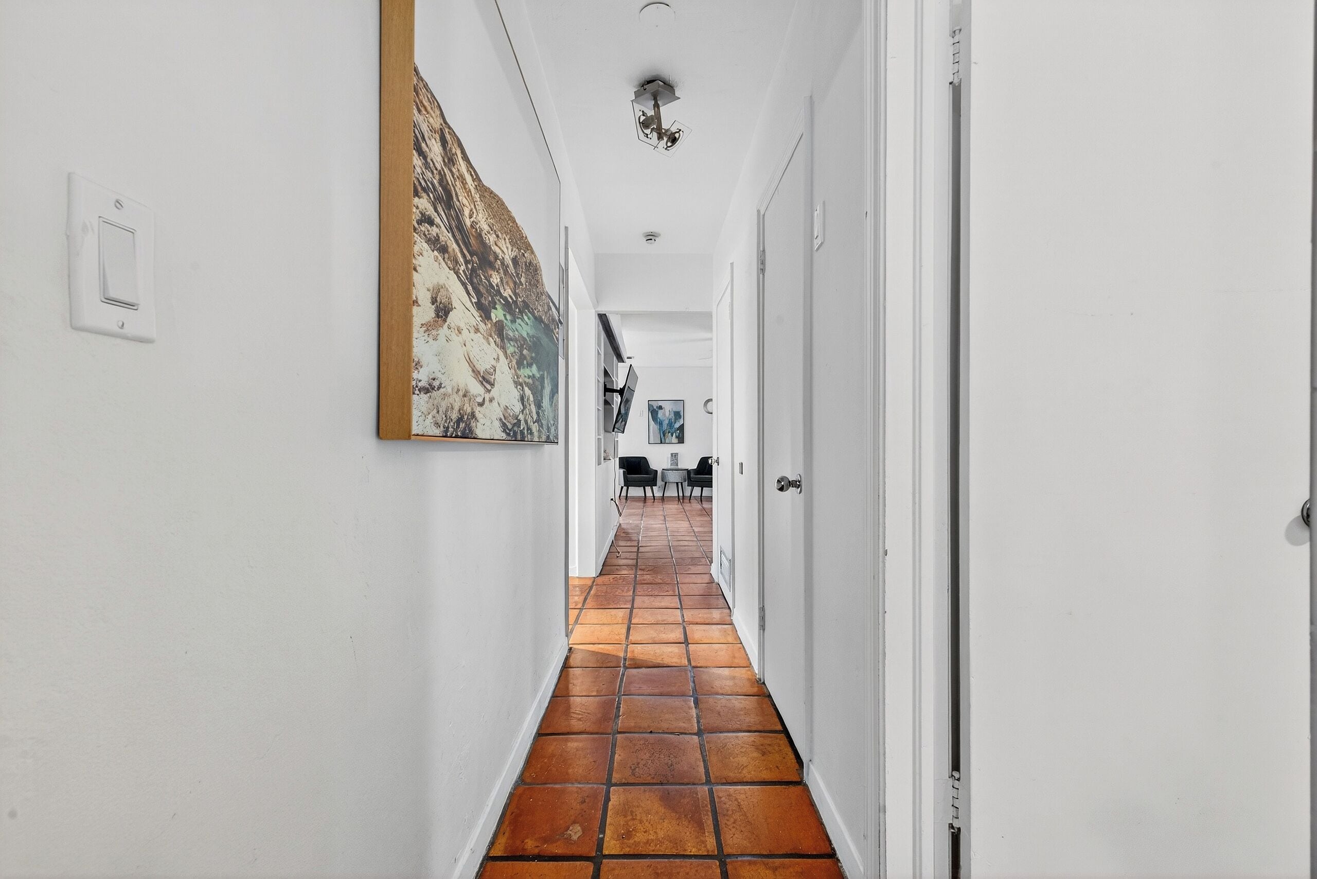A hallway with white walls, a tiled floor, and a large abstract painting on the left.