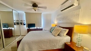 A bedroom with a bed, bedside lamps, a ceiling fan, an air conditioner, a TV, and a mirror.