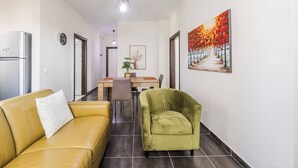 Un salon moderne avec un canapé jaune, un fauteuil vert, une table à manger avec des chaises, un réfrigérateur, une horloge et un tableau représentant un chemin bordé d’arbres.