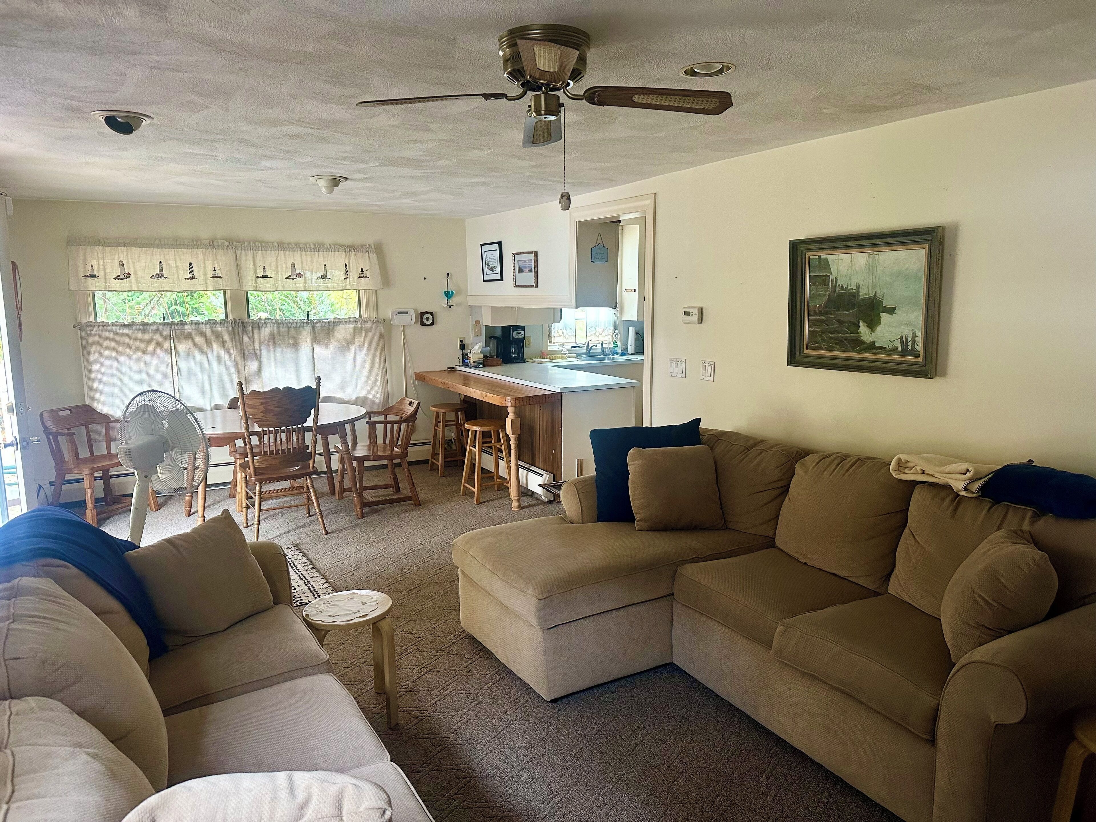 Ein Wohnzimmer mit Ecksofa, einem Esstisch mit Stühlen, einer Küchenzeile und einem Deckenventilator.