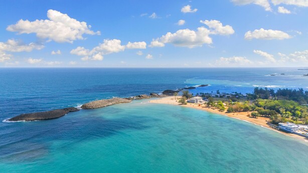 Vista costeira com águas turquesa translúcidas, uma praia arenosa e um pequeno povoado com edifícios e vegetação.