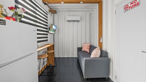 En kompakt opholdsstue med et tekøkken, en grå sofa med pink puder, et lille træbord og et vægmonteret TV.