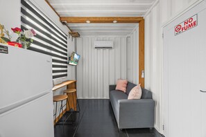A compact living space with a kitchenette, a gray sofa with pink cushions, a small wooden table, and a mounted TV.