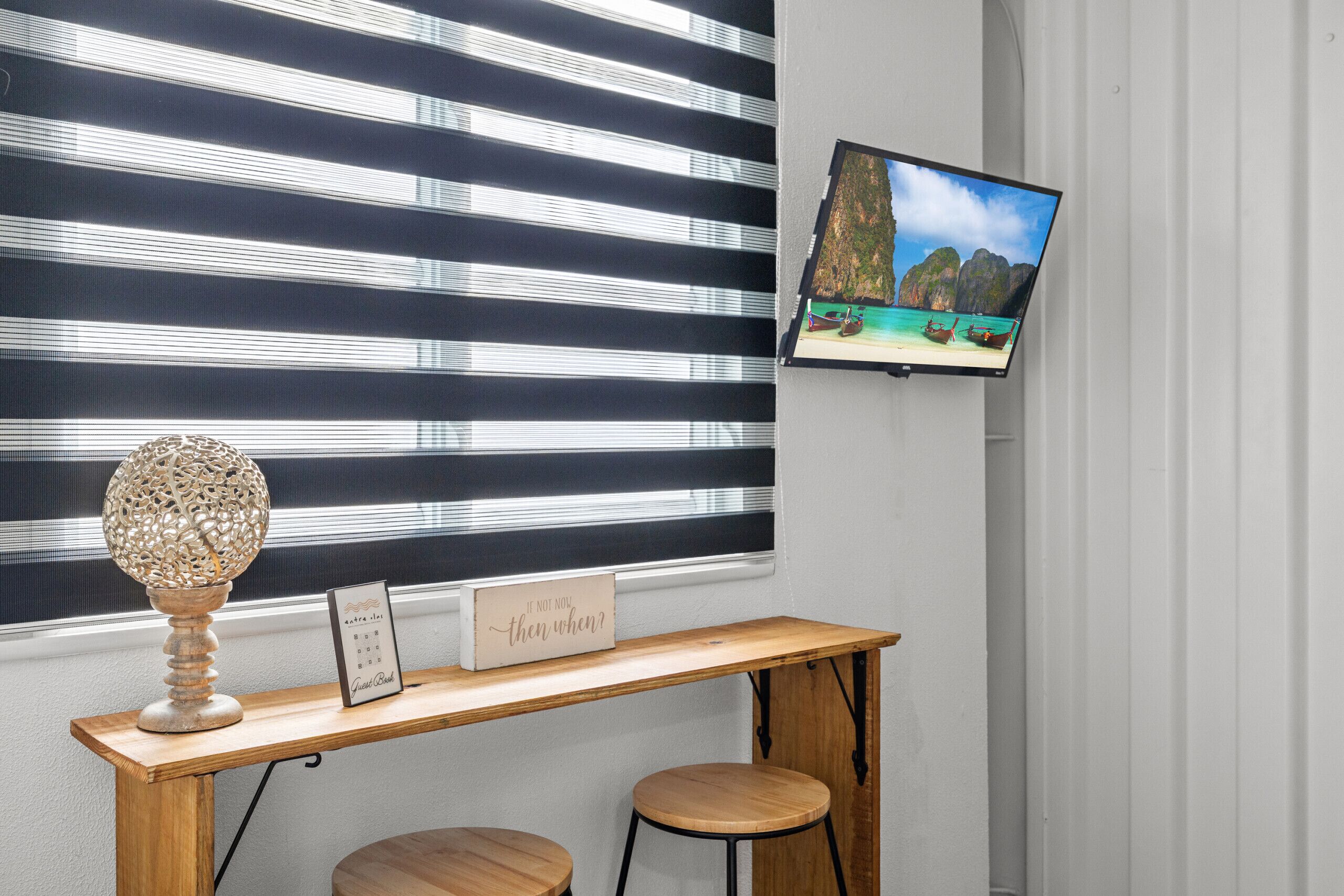 Una habitación con pared a rayas, una mesa de madera con dos taburetes, una lámpara decorativa, un letrero enmarcado y un televisor que muestra una imagen panorámica.