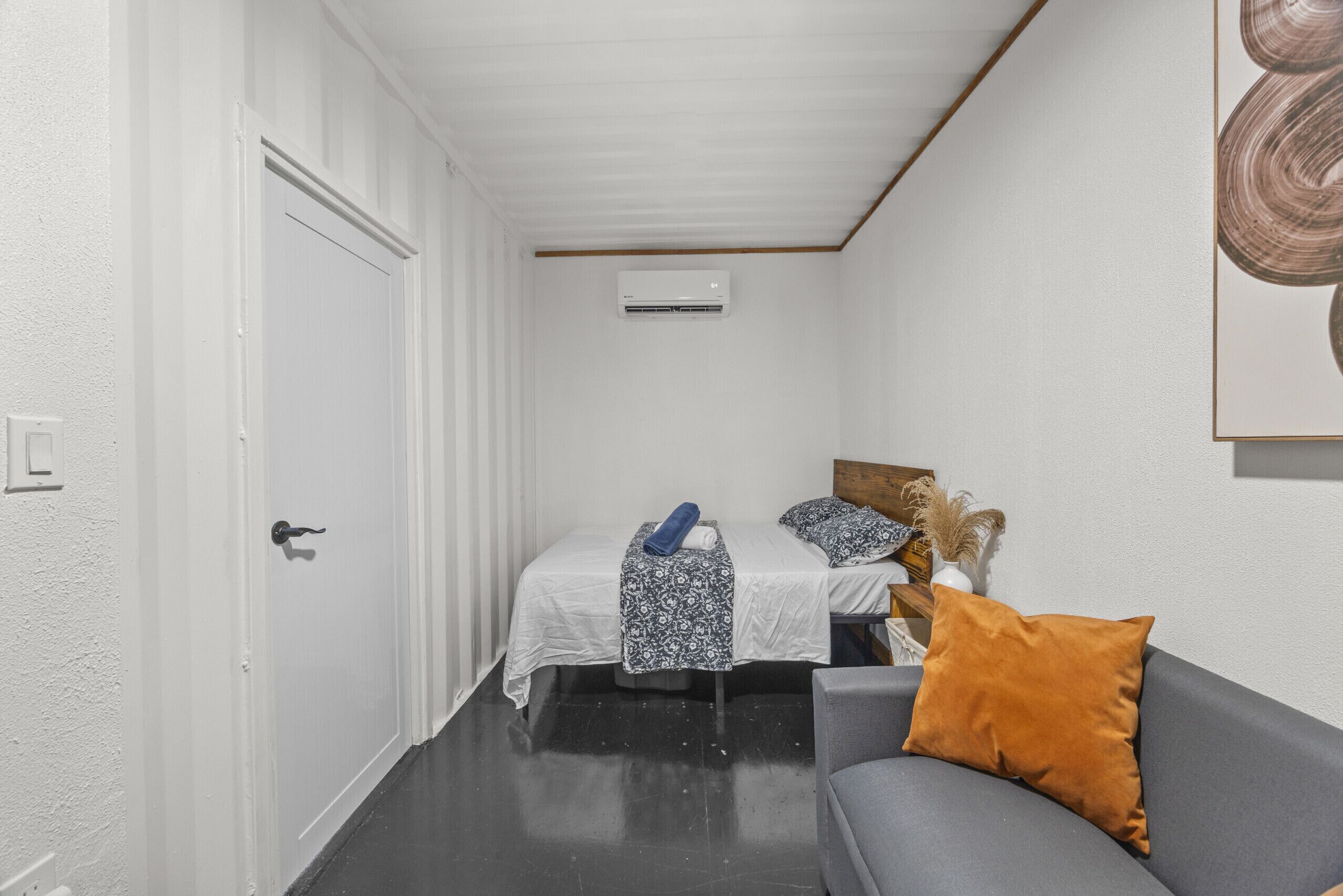 Un pasillo estrecho que conduce a un dormitorio con cama, un sofá gris y un equipo de aire acondicionado en la pared.