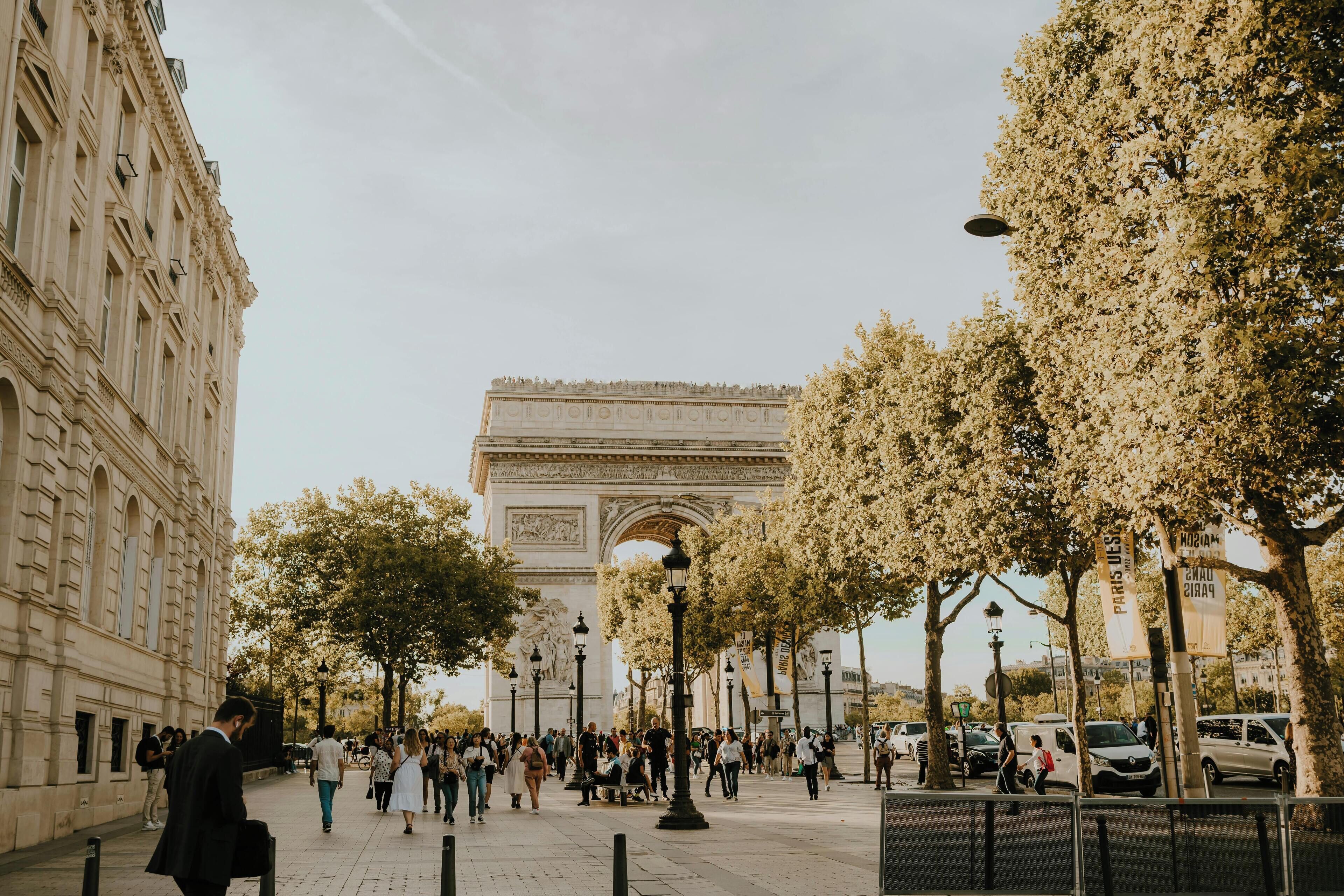 A wide pedestrian area with a large historical arch in the background, surrounded by trees and street lamps.