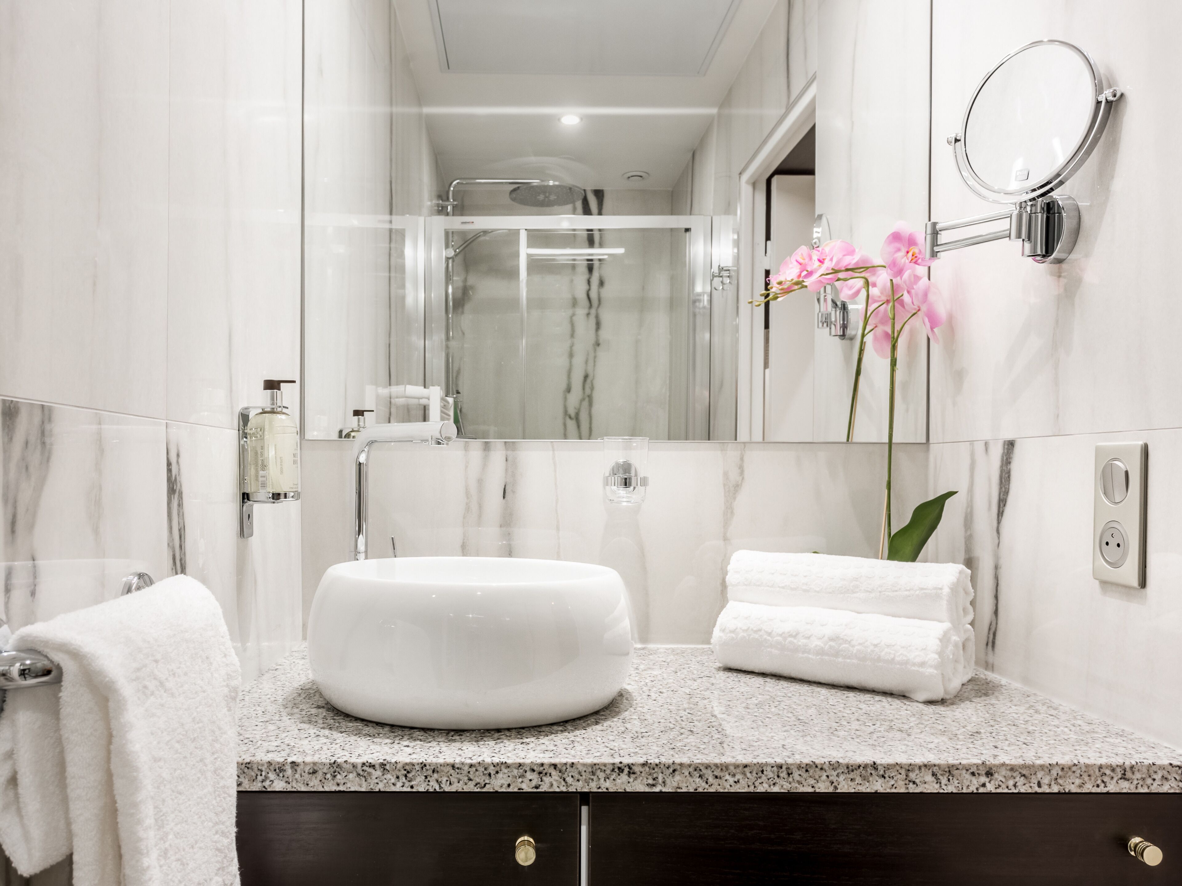 A modern bathroom with a large oval sink, a glass shower enclosure, and a wall-mounted mirror with a pink orchid.