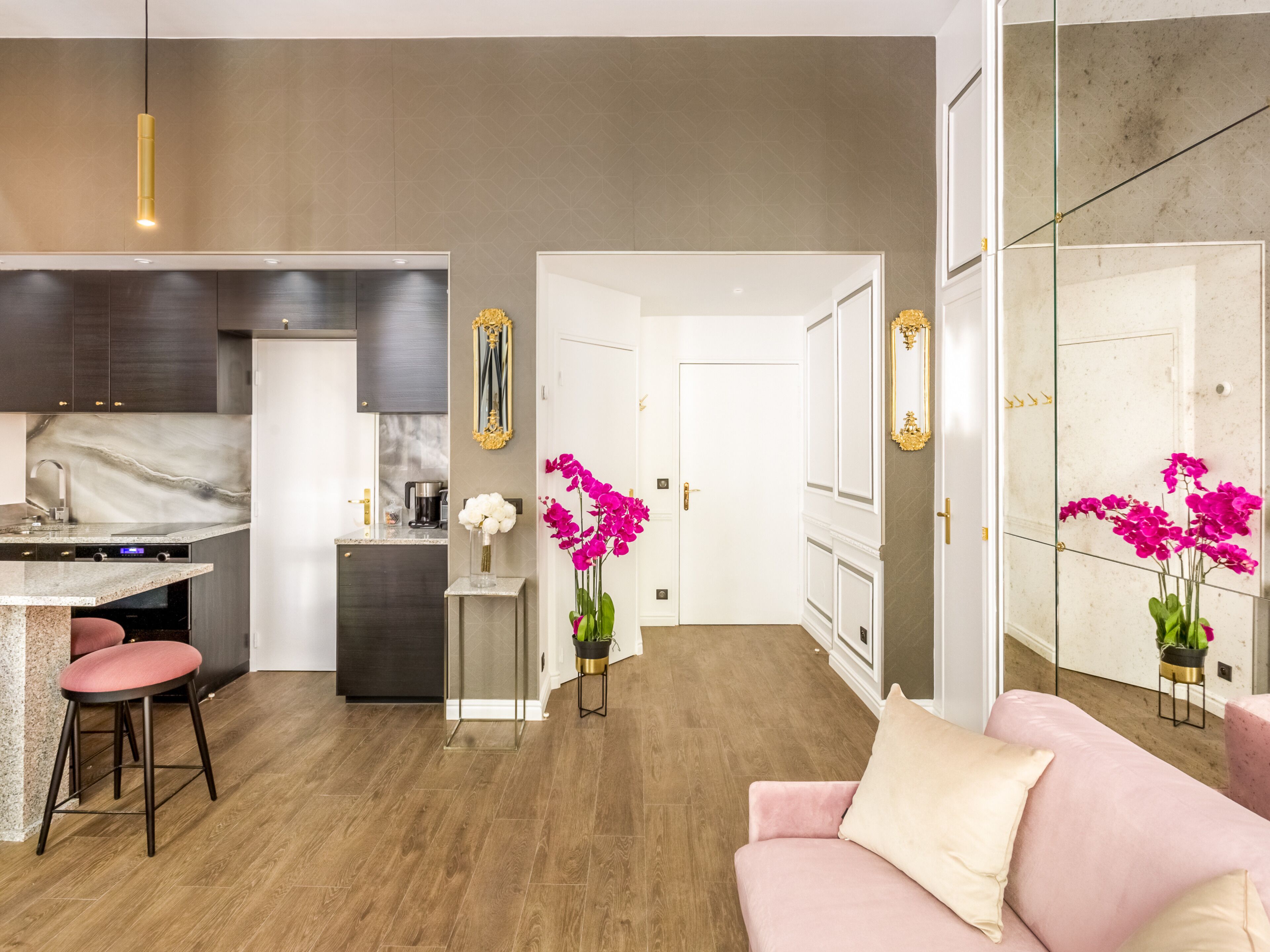 A modern kitchen with dark cabinets, a marble countertop, and a built-in oven. There is a pink sofa and a small table with a vase of flowers in the living area.