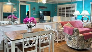 Un comedor con mesa de madera y sillas blancas, una cocina con paredes verde azulado y una sala de estar con un sillón estampado y un sofá rojo.