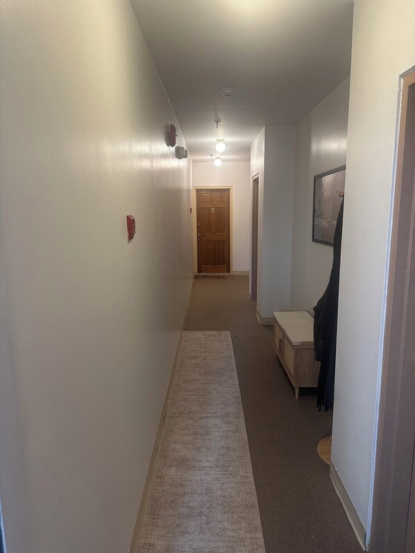 A narrow hallway with a carpet, a wooden door at the end, and a bench with a coat hanging on it.