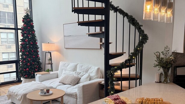 Una sala moderna con un sofá blanco, una mesa de centro y un árbol de Navidad. Se observa una escalera de diseño espiral y una cocina con mostrador.