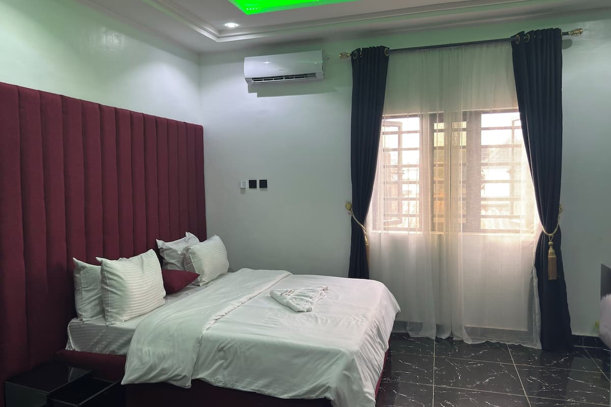 Quarto de hotel com cama, cabeceira vermelha, roupa de cama branca, piso de azulejos pretos e uma janela com cortinas.