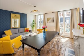 Un salon moderne avec un canapé, des fauteuils jaunes, une table à manger et une grande fenêtre donnant sur des immeubles.