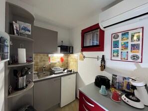 A compact kitchen with a sink, microwave, and a coffee machine. There are shelves with various items and a window with a red frame. - The Little Arbanais : Ideal studio near the sea (Hyères)