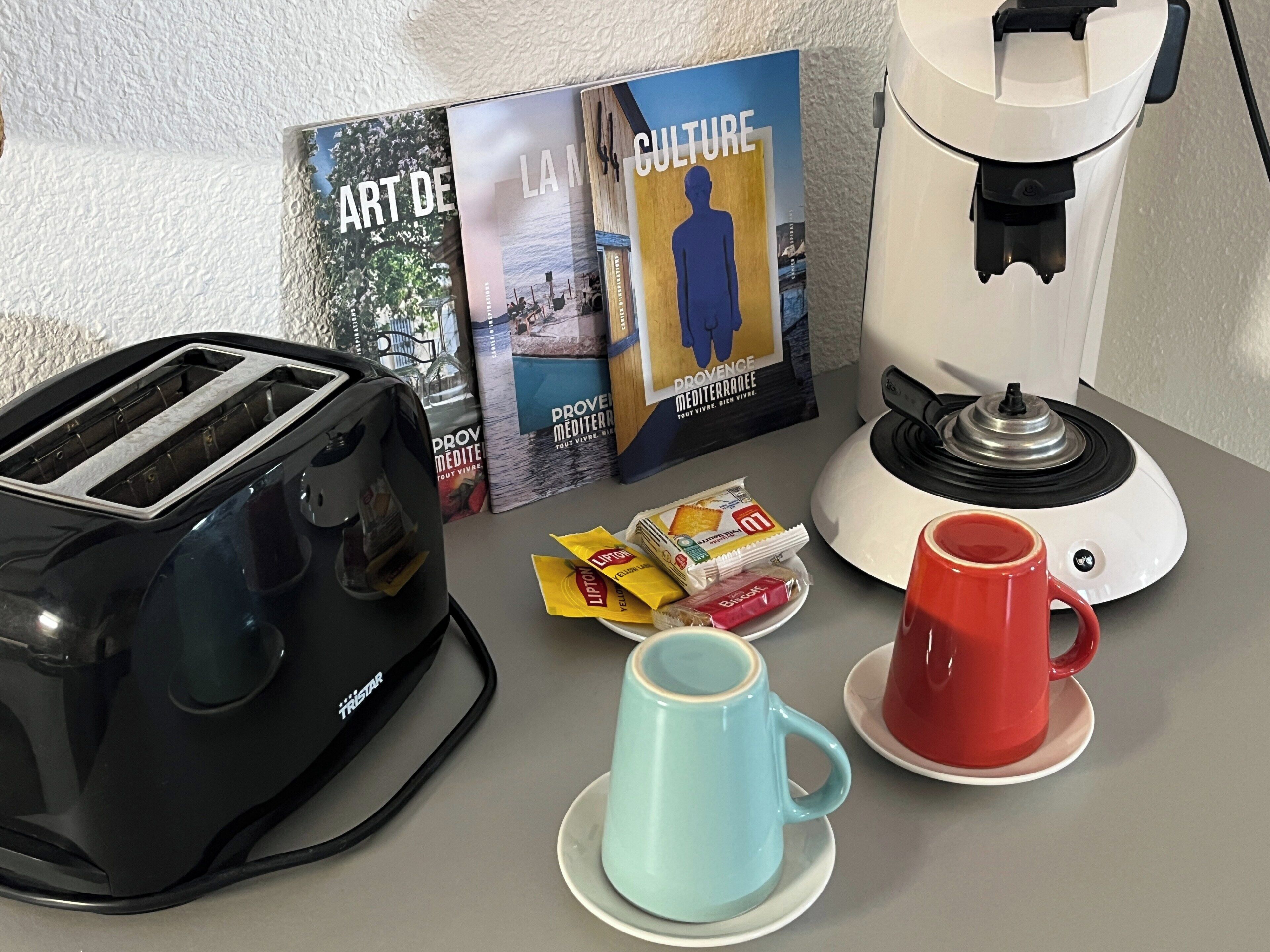 A kitchen counter with a toaster, a coffee maker, a red mug, a blue mug, and some packaged snacks.