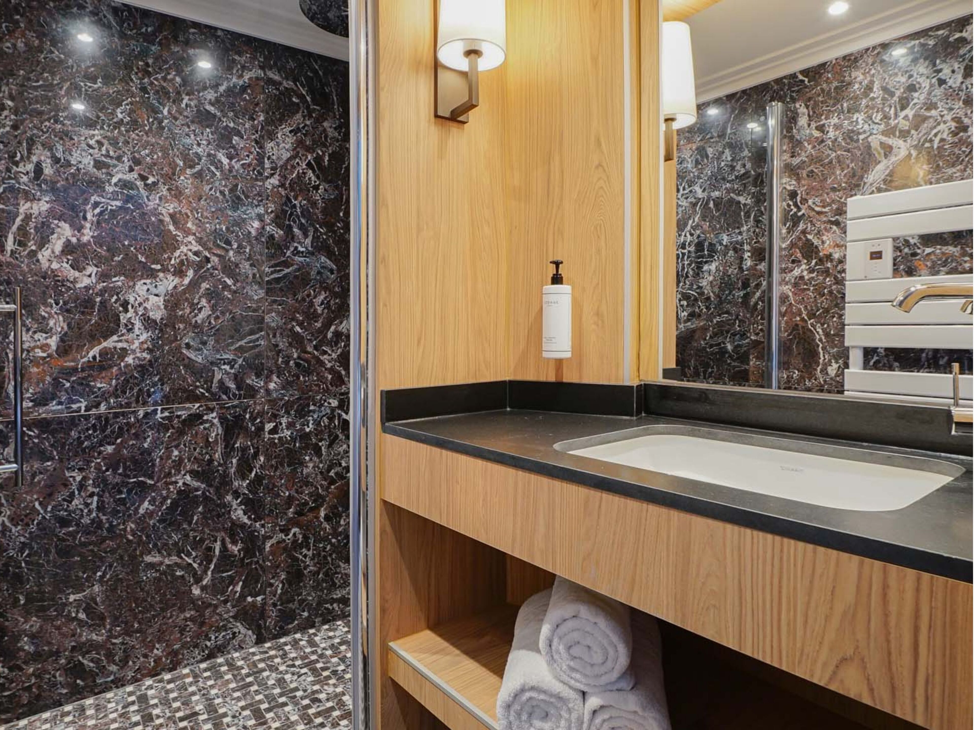 Un bagno con parete in marmo, mobile lavabo in legno, lavandino, porta asciugamani e specchio.
