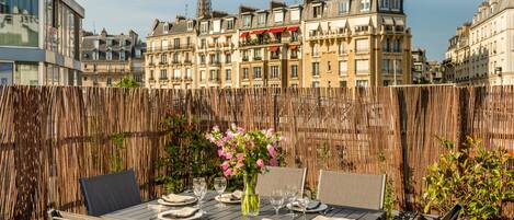 An outdoor dining area with a table set for four, surrounded by chairs, and a view of a cityscape with buildings and a tower in the background.