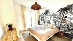A bedroom with a bed, a wooden table, a small stool, a woven hanging light, and a wall mural of palm trees. - Casa Alegria (Tunes)