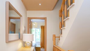 Ein Flur mit einer Holztreppe, einem Spiegel, einer Lampe und Blick in einen Raum mit Bett und Vorhängen.