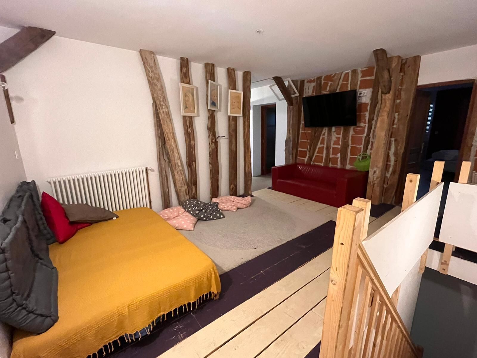 Un dormitorio con cama, sofá rojo, vigas de madera, televisor y radiador.
