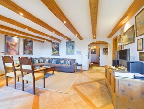 Ein großzügiger Wohnbereich mit einer Couch, Holzstühlen, einem Couchtisch und einem Fernseher. An den Wänden befinden sich gerahmte Bilder und es gibt eine Holzbalkendecke.