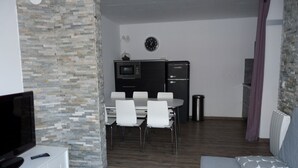 Un espacio habitable compacto con televisor, una mesa de comedor con sillas, una kitchenette y un sofá con almohadón.
