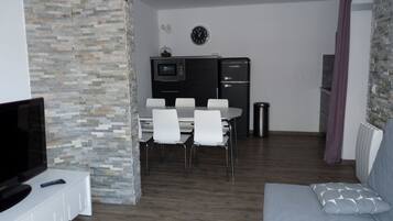 Un espace de vie compact comprenant un téléviseur, une table à manger avec des chaises, un coin cuisine et un canapé avec un coussin.