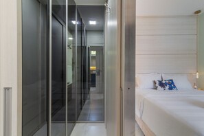 Una habitación de hotel moderna con un separador de vidrio, una cama bien hecha con ropa blanca y un cojín decorativo, y un baño visible al fondo.