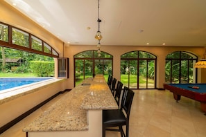 A spacious indoor area with a long granite countertop, a pool table, and large arched windows offering a view of the outdoors. - Casa Zafiro 7 Bdrs Private Pool in Los Suenos (Jacó)