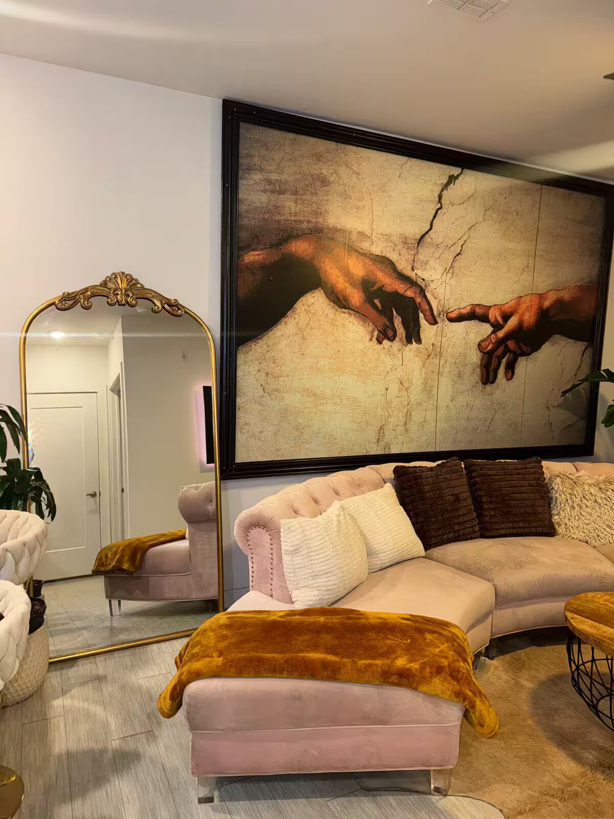 A living room with a large painting of hands reaching towards each other, a tufted sofa with cushions, a gold-framed mirror, and a small round table.
