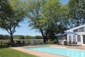Ein Swimmingpool mit Liegestühlen und Sonnenschirmen, umgeben von Bäumen und einem weißen Gebäude mit großen Fenstern.