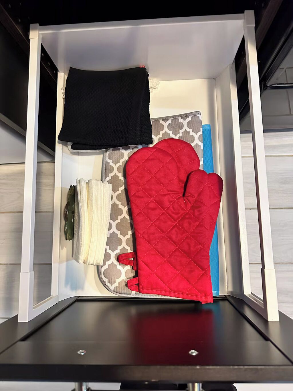 A kitchen drawer with a black towel, a red quilted pot holder, a white cloth, and a patterned cloth.