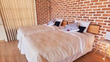 A hotel room with a brick wall, two beds with white bedding, wooden flooring, and a window with curtains.