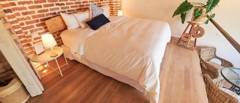 Un dormitorio con pared de ladrillo, cama con ropa de cama blanca, una mesita con lámpara y mobiliario de mimbre.
