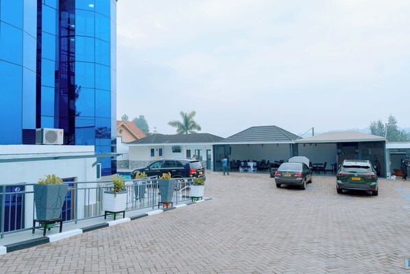 A parking area with two cars, a building with blue glass facade, and a covered outdoor seating area. - Dorlan Hotel (Kigali)
