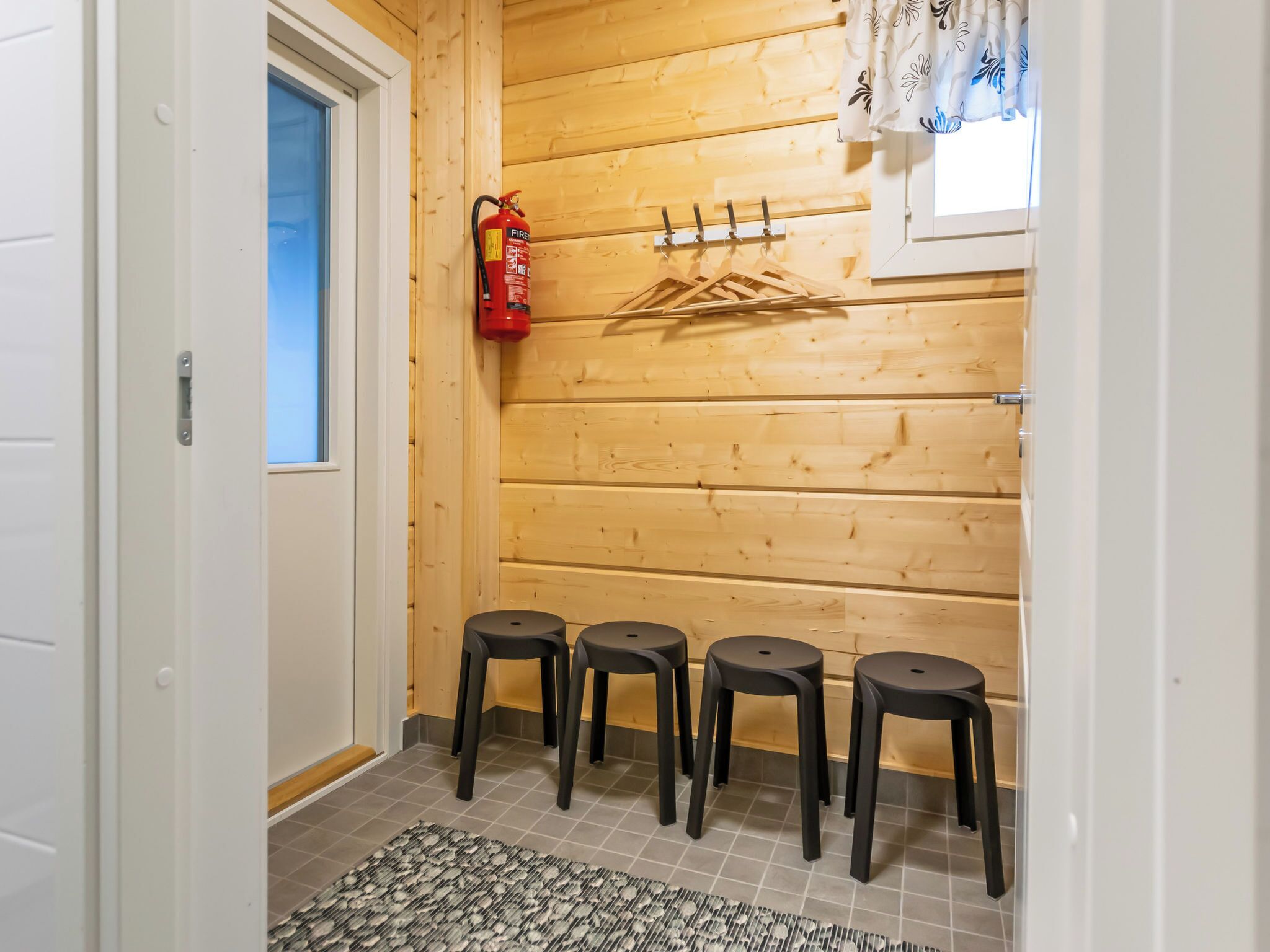 Un couloir aux murs en bois, un extincteur, un porte-manteau et quatre tabourets noirs.