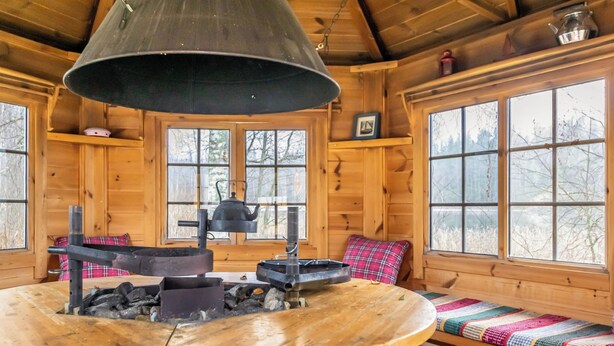 A cozy cabin interior with a central wood-burning stove, a hanging light fixture, and a bench with a colorful blanket. - Joutsenranta by Interhome (Tuusniemi)