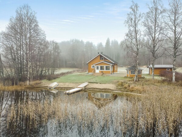 Een rustieke hut met een groot raam, omgeven door een vijver en dichte bomen.